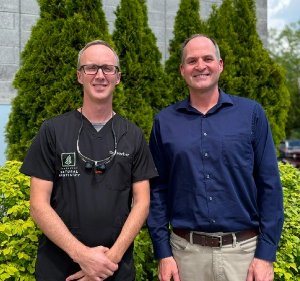 Two smiling men stand side-by-side outdoors in front of green bushes. The man on the left wears black dental scrubs embroidered with "Dr. Harker" and a dental logo, with loupes hanging around his neck. The man on the right wears a dark blue button-down shirt.